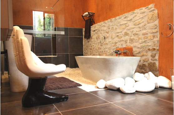 Salle de bain design avec baignoire balnéo devant murs en pierre entouré d'un enduit orange finition mat. Carrelage grands marron dans douche italienne et sol 