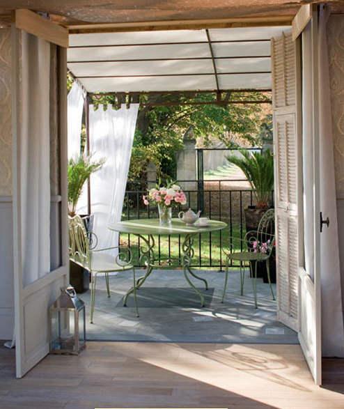 Aménagement d'une terrasse déco intime dans le prolongement du salon avec toit verrière et rideaux brise vue accrochés à la structure en fer forgé 