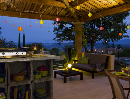 cuisine d'été extérieur aménagée sous auvent en bois, grande dalle de ciment au sol, salon de jardin en rotin, luminaire lampion de couleurs