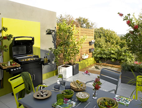 une cuisine d'ete d'exterieur amenagee sur une terrasse. grand barbecue multi-fonctions mobilier outdoor metal couleur gris et vert, jardinière bac a plante carre, couleur vert et blanc