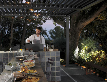 Une cuisine d'été outdoor aux lignes épurés et tons de gris aménagée sous pergola. Bloc cuisson et bar en maçonnerie avec carrelage 15X15 gris