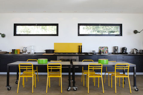 Dans une cuisine ouverte la couleur jaune de la crédence en verre et les chaises métalliques se conjuguent de façon très déco avec les meubles noir et la table industrielle