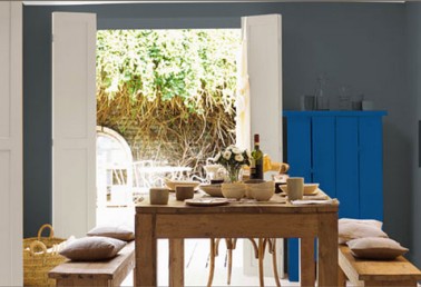 decoration-salle-a-manger-peinture-murale-gris-soutenu-et-gris-clair-meuble-peint-bleu-intense-associe-a-table-et-banc-hetre-naturel-coussins-couleur-lin decoration salle à manger minimaliste avec ensemble table et banc en hêtre naturel coussins en lin, peinture murale gris soutenu associé a un bleu electrique sur une armoire peinte.