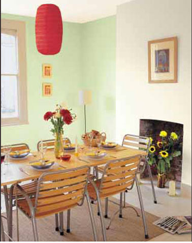 Peinture murale vert amande pour une salle à manger associée à sa couleur complémentaire, le rouge pour le luminaire. Le mur de la cheminée peint en blanc cassé renforce l'atmosphère pleine de fraicheur de cette pièce à vivre.