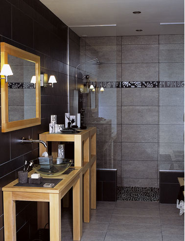 Dans cette salle de bains, un jeu de formes et de lignes en gris et noir. Pour la douche à l'italienne, un carrelage mural gris réhaussé par une frise horizontale en noir et gris brillant. Le sol de la douche est réalisé en galets gris joints noir. Les meubles en bois reprennent le jeu des formes rectangulaires du carrelage mural