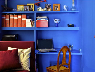 Espace bureau créé dans un salon avec étagère et mur peint en bleu intense. chaise et accessoires de bureau en orange pour créer un contraste fort et chaleureux