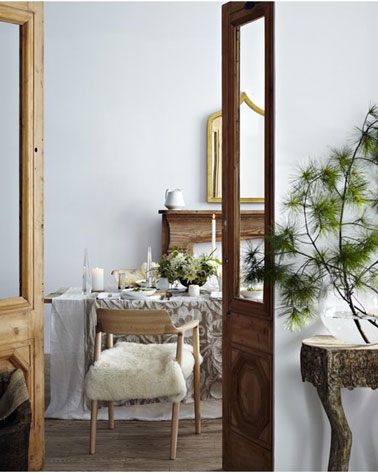 Sur la table de Noël d'une salle à manger à la déco zen, des nappes en lin et coton en superposition mixant nappe blanche et lin décorée de bougies blanches et de branchages pour la touche couleur