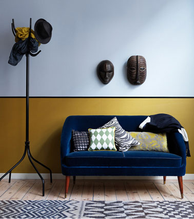 Salon design africain avec murs peinture couleur bleu, soubassement peinture jaune, ligne peinture noir, coussins tissu africain sur petit canapé bleu Monaco