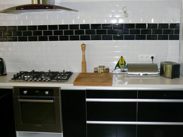 Décoration de cuisine en noir et blanc. Meubles bas en stratifié noir brillant, plan de travail stratifié beige. Murs en crédence en carrelage blanc rehaussé d'un Listel noir 3 x 15 cm. 
