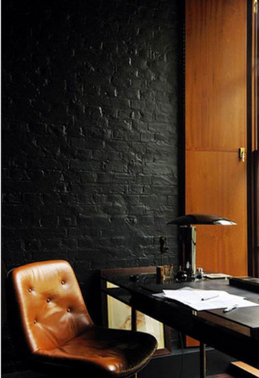 Coin bureau dans salon design. Le mur de brique est peint en noir brillant et met en valeur le cuir du fauteuil et les meubles chêne doré