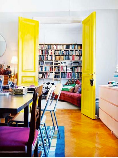 Entre salon et salle à manger, porte peinte en jaune vif, murs blanc, coussins chaises violet, canapé rouge