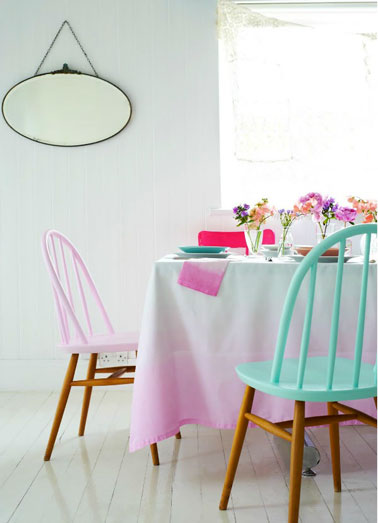 Ambiance printanière dans une salle à manger blanche apportée par les couleurs pastel utilisées pour repeindre l'assise et le dossier des chaises en bois et l’explosion des couleurs utilisées pour la décoration de la table.