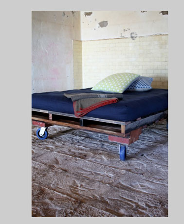 Lit palette monté sur roulettes dans une chambre d'adulte. les palettes du sommier sont laissées brutes pour s'intégrer au style épuré de la chambre