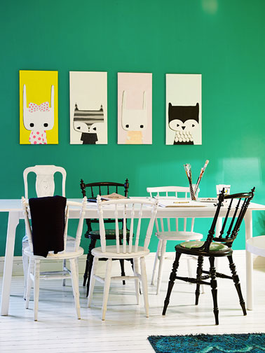 Salle à manger avec chaises en bois dépareillées. Deux chaises peintes en noir brillant contrastent avec le blanc pur de la table et des autres chaises. L'ensemble est mis en valeur avec la couleur vert émeraude de la peinture murale