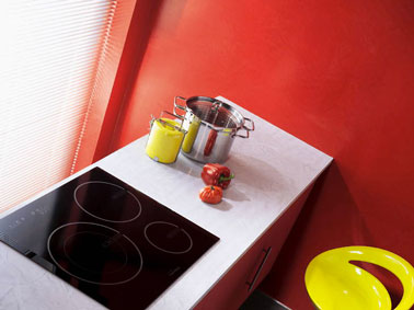 Façades meubles bas de l'espace cuisson d'une petite cuisine peints en rouge vif, sur les murs une peinture effet béton couleur rouge en rappel, table à induction noir, plan de travail blanc apportent un contraste fort. Tabouret de bar jaune vif.