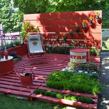 Sol terrasse en palette bois peint en rose fushia Sol terrasse bois en palettes aménagé dans un coin du jardin. sur les 6 palettes peintes en rose fushia, transat et bidon de recup formant le salon de jardin sont repeints dans la même couleur. Sur la claustra bois fermant l’espace, des jardinières en parpaing assorties