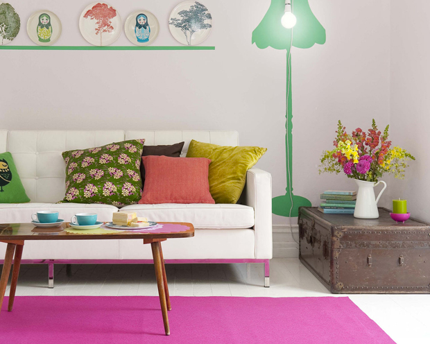 Couleur pour agrandir un salon. Les couleurs fushia, vert jade et jaune permettent d'agrandir l'impression d'espace de ce petit salon. Peinture murale gris pastel, tapis rose fushia, pochoir lampadaire vert jade sur le mur, bouquet de fleurs, frises et coussins reprennent ces couleurs vives en touches