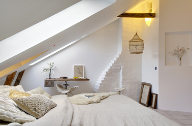 Une chambre beige et blanche aménagée dans un ancien atelier d'artisan. Pour construire une décoration épurée, les murs blanchis soulignent les poutres du plafond, le linge de lit beige renforce la douceur ambiante.