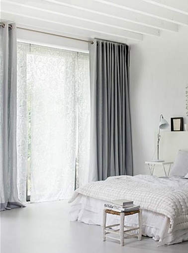 Déco chambre grise et blanc dans laquelle la seule touche de gris est apportée avec la couleur gris souris des double-rideaux. au dessus du lit blanc petit cadre photo noir. Pied de lit, tabouret bois