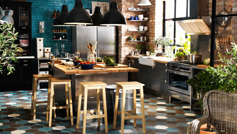 Une déco de grande cuisine qui mixe meubles noirs et inox, ilôt central inox plateau bois clair grandes suspensions noires et un carrelage sol qui rappelle l'ensemble des couleurs de la cuisine