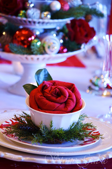 Serviette rouge pliée en forme de fleur posée dans coupelle blanche et décorée de feuillage vert