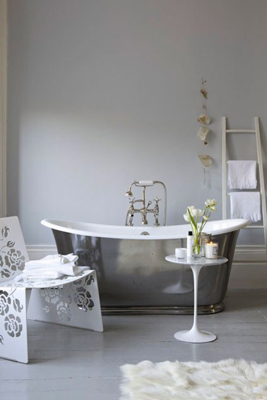 Salle de bain grise et blanc avec baignoire îlot finition étain, échelle en bois clair qui fait office de porte serviette posé sur mur gris pastel. 