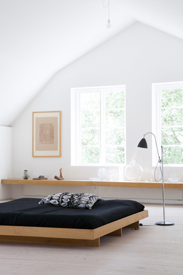 Pour la déco de cette chambre zen, juste un grand lit confortable en chêne clair recouvert d'un dessus lit noir pour contraster avec la pureté du blanc des murs et une longue étagère en chêne clair également. Une déco sobre pour une ambiance pleine de sérénité