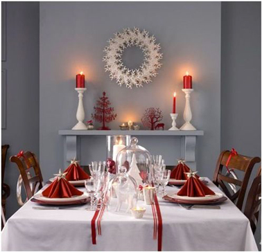 Déco table de Noël rouge et blanc dans une salle à manger grise : Nappe blanche, chemin de table ruban rouge, serviettes rouge, sur la cheminée chandelier céramique blanche et bougies rouges