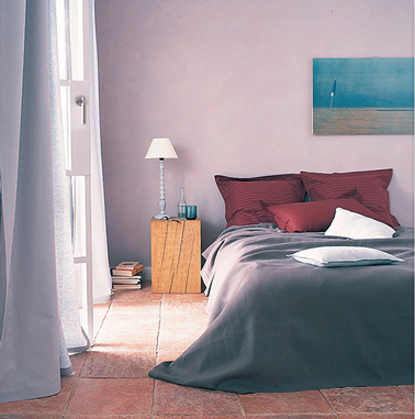 Ambiance cosy dans une chambre parentale avec une harmonie de teintes douces. Aux murs une subtile teinte de peinture gris au reflet rosé, linge de lit et rideaux brun taupe relevé par la couleur des oreillers rouge velouté