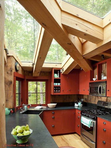Sublime cuisine rouge et plan de travail gris anthracite installée dans une véranda face au jardin avec de grosses poutres bois et un toit en verre