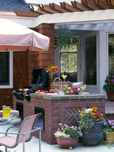 Cuisine dété aménagée près de la maison avec un bloc en briques rouge pour intégrer évier et barbecue. Plan de travail en carreaux gris imitation béton