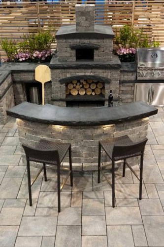 Une cuisine extérieure tout confort installée sous une pergola. Construction en pierre, plan de travail en ardoise avec four à pizza avec niche pour réserve bois et barbecue inox