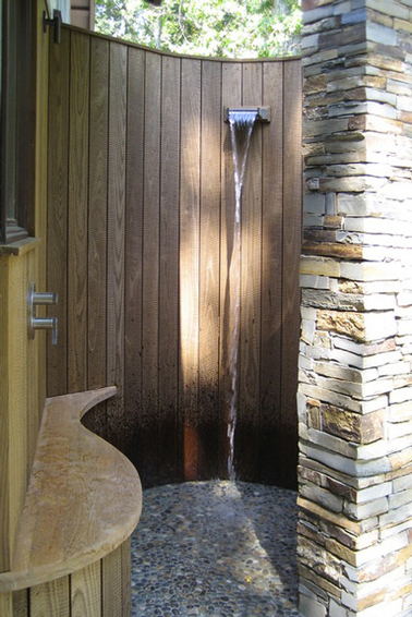 Une douche de jardin installée dans un patio construit en arrondi avec des planches de teck et un pilier de pierres pour terminer l'arrondi