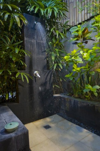 Douche de jardin installée dans un havre de végétation et de pierres noires. Au sol un carrelage de grands carreaux de pierre beige