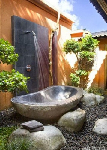 Une douche d'extérieure avec une baignoire en pierres pour receveur  installée dans un environnement aux couleurs magiques et un sol de gros galets.