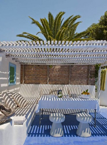 Superbe ambiance sous cette pergola bois d'une maison grec. Sur le sol en ciment blanc est peint un grand tapis bleu qui sublime le blanc des banquettes et de la pergola. 