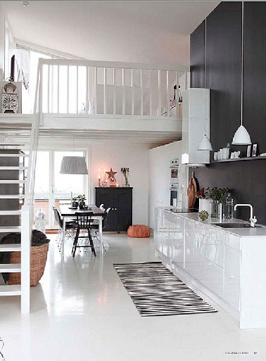 Dans un espace ouvert sur le salon salle à manger, une cuisine blanche mise en valeur par un mur et crédence noir qui fait ressortir l'aspect ultra brillant des façades des meubles