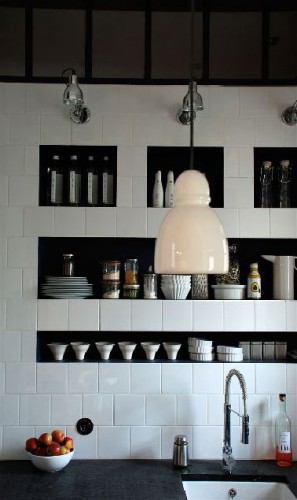 Dans le mur en carrelage blanc au dessus du plan de travail d'une petite cuisine, des niches peintes en noir pour le rangement de la vaisselle blanche, une solution esthétique et pratique pour gagner de la place 