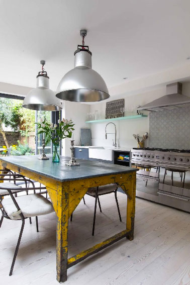 Dans cette cuisine, la crédence se pare de motifs rétro gris se mariant parfaitement avec l’inox omniprésent dans la pièce. La  table en acier brut d’origine industrielle offre une touche de couleur jaune bouton d’or éclatant qui dynamise la dominante de blanc et gris.