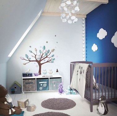 Un mur bleu nuit, des stickers et du mauve pour une chambre bébé fille de rêve Un mur peint en bleu nuit vient sublimer cette chambre bébé fille mansardée. Et pour un univers encore plus remplit de rêves, des formes nuages sont disposées sur le mur ou encore sur le lustre. Des stickers muraux en relief viennent quant à eux créer de toute pièce un monde imaginaire et créatif fait d’arbres, d’étoiles et de chouette. Des notes de mauves toutes en nuance viennent égayer la pièce en la féminisant. La chambre parfaite pour les petites princesses rêveuses !