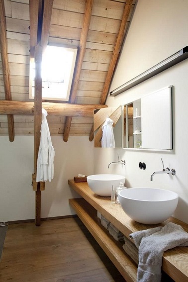 Aménagement d’une petite salle de bain sous les combles qui valorise la structure bois d’origine pour créer son ambiance chaleureuse. Poutres apparentes, plafond lambris laissés brut et traités contre l’humidité et le long meuble vasque en bois accueille 2 grandes vasques rondes. 
