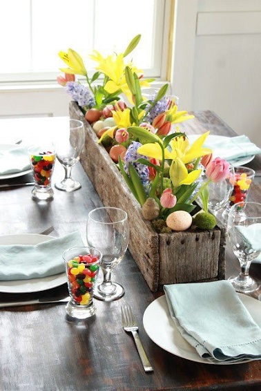 Un centre de table fleuri pour une déco raffinée et printanière, parfaite pour Pâques. Très simple et si charmante cette déco de table printanière ! Le centre de table garni et fleuri constitue à lui seul la déco de cette table. Dans un bac à fleurs en bois rectangulaire ont été plantés des fleurs roses, mauves et jaunes dont les couleurs joyeuses viennent réveiller cette table à la base sobrement décorée. Quelques œufs cachés dans la jardinière déco entre la verdure rappellent le thème pascal en subtilité. Et parce que Pâques est aussi un véritable moment de gourmandise, des verres remplis de bonbons multicolores complètent la déco autour des couverts. Une table de Pâques pleine d’idée et facile à reproduire chez-soi, on adore ! Photo : Trouvée sur mycraftilyeverafter.com / Migonis Home