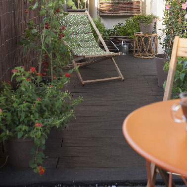 Petite terrasse aménagée avec des dalles en bois composite fermée par de la brande et décorée de plantes et meubles de jardin en bois.
