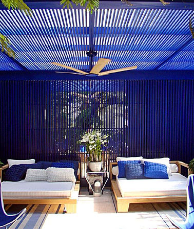 Abritée du soleil et du vent sous un patio construit en bois peint en bleu, une terrasse design aménagée avec de larges bains bain de soleil en bois et toile blanche.