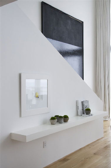 Une déco couloir d’exception dans une maison moderne, avec une montée d’escalier dessinant une diagonale laissant apparaitre une immense toile noire semblant sortir des murs peints en blanc.