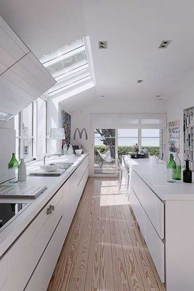 Lumineuse avec ses vélux et baies vitrées une deco de cuisine aménagée en longueur avec des meubles laqué blanc posés en vis à vis sur un parquet clair. Une ambiance désign et épurée.