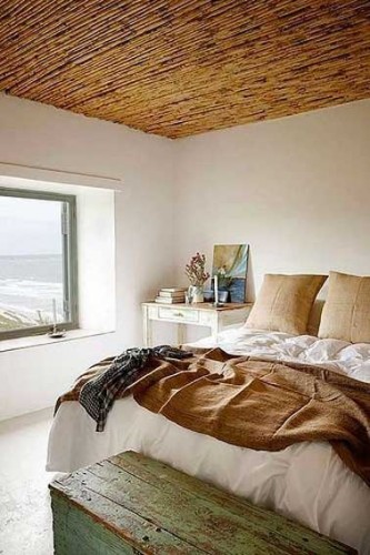 Ambiance cocooning dans une chambre parentale décorée en dégradé de couleur lin et teintes naturelles sous un plafond en lambris verni et un coffre en bois en pied de lit.