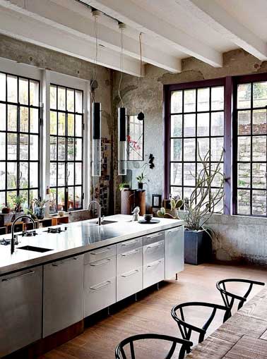 Beau mix de style avec le bois brut de la table et celui des verrières peint en violet. Déco contrastée par l’îlot central en acier et les luminaires design