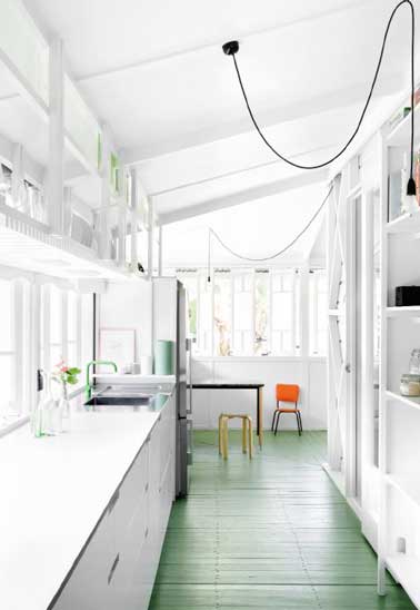 Cuisine en longueur avec une verrière posée tout le long de la pièce. Sol peint en vert, chaise orange et tabouret jaune colorent la déco de cette cuisine blanche. 