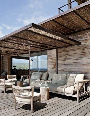 Une pergola canisse ombrage une terrasse en bois d’une maison Corse. Le mobilier de jardin en teck contemporain et confortable à la fois avec ses grands coussins épais invite à la détente. 